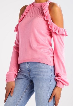 Žena oblečená v světlých modrých džínách a růžovém svetru s odhalenými rameny a volánky podél vykrojení na ramenou.