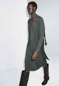 Szara sukienka z długimi rękawami i dekoltem w kształcie V, z drapowaniem i bocznymi rozcięciami, w połączeniu z ciemnobrązowymi kozakami do kolan. Gładka faktura materiału.