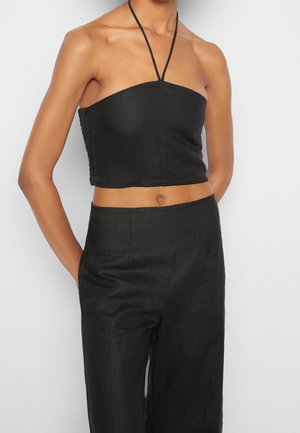 Kvinde iført sort ærmeløs halter crop top kombineret med sorte bukser med høj talje, hænderne i lommerne, foran en ensfarvet hvid baggrund.