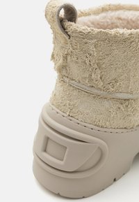 Botine bej cu un superior texturat și pufoasă și o talpă solidă din cauciuc. Prezintă o formă rotunjită și o buclă de prindere pentru o îmbrăcare ușoară.
