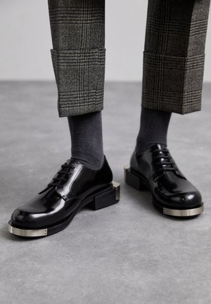 Scarpe oxford nere lucide con punta quadrata, con accento argentato sulla suola, abbinate a pantaloni grigi con fantasia e calzini grigi.