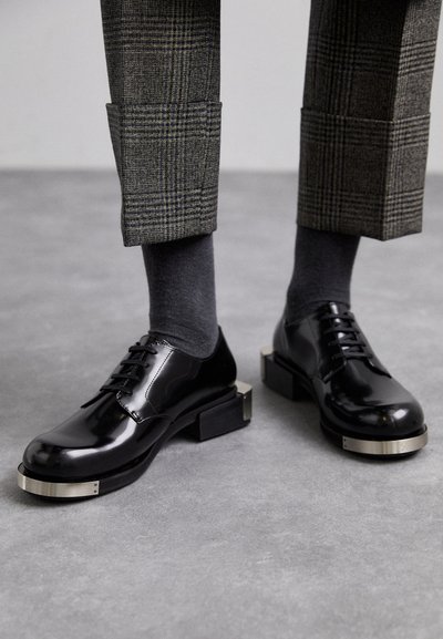 Zwart gepolijste oxford schoenen met vierkante neus, zilveren accent op de zool, gecombineerd met grijs met patroon broeken en grijze sokken.