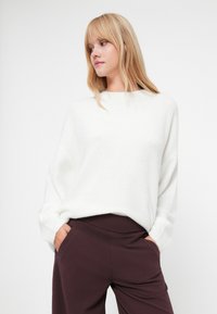Suéter blanco y esponjoso de corte holgado y mangas largas, combinado con pantalones burdeos oscuros con bolsillos laterales. Diseño minimalista.