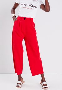 Pantalon rouge à jambes larges avec une fermeture à boutons et des plis avant, associé à un t-shirt blanc avec un motif graphique et des sandales blanches à enfiler. Texture lisse.