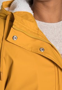 Żółta wodoodporna kurtka z wysokim kołnierzem, podszyta polarem, z srebrnymi zamkami błyskawicznymi i zatrzaskami do zapinania.