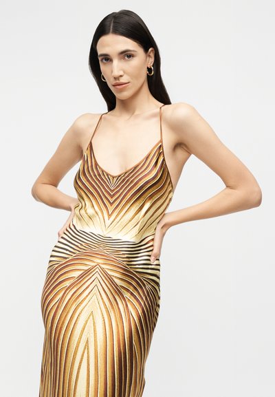 Γυναίκα με μακριά σκοτεινά μαλλιά φοράει αμάνικο φόρεμα με χρυσά και καφέ γεωμετρικά μοτίβα, ποζάροντας με τα χέρια στη μέση.