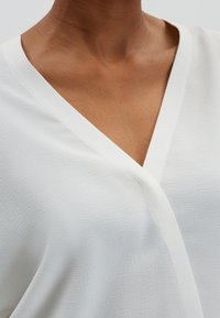 Blusa bianca avvolgente con scollatura a V, tessuto testurizzato e dettagli di cucitura puliti. Inquadratura sul collo e sulla zona del busto.