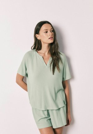 Kvinde med langt brunt hår iført en lys grøn kortærmet top og matchende shorts, stående op ad en ensfarvet, lys væg.