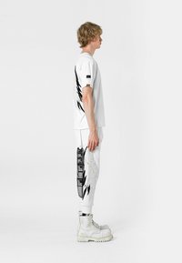 Camiseta blanca con un diseño de alas negras en la parte posterior, combinada con pantalones blancos que tienen gráficos laterales y acentos negros; llevada con botas blancas robustas.