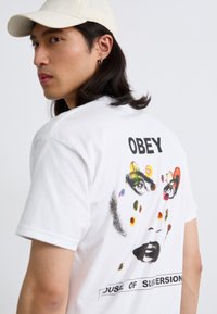 Bela bomba majica iz 100% bombaža s barvitim grafičnim umetniškim motivom obraza na hrbtu, z besedilom "OBEY" in podnapisom "HOUSE OF SUBVERSION".