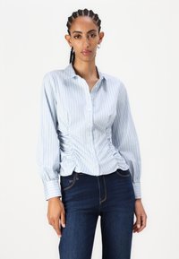 Lys blå skjorte med lange ermer og knapper, hvite striper, samlet sider, og krage, kombinert med mørke jeans. Glatt stofftekstur.