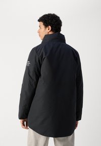 Chaqueta exterior negra con un cuello alto, mangas largas y un ajuste ligeramente holgado. Presenta una tela texturizada y un detalle de marca minimalista en el hombro.