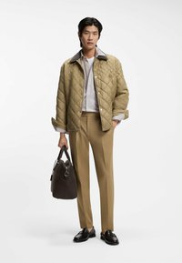 Beige tikattu takki, jossa on tumma kaulus, vaalea paita, vaaleanruskeat housut ja ruskea laukku. Malli seisoo kädet takin taskuissa, jalassa mustat loaferit.