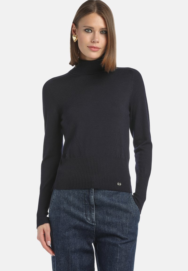 Marinefarbener Rollkragenpullover mit geripptem Saum und Bündchen, aus einem weichen Material gefertigt. Mit einem kleinen Logodetail an der unteren linken Seite. Kombiniert mit blauen Jeans.