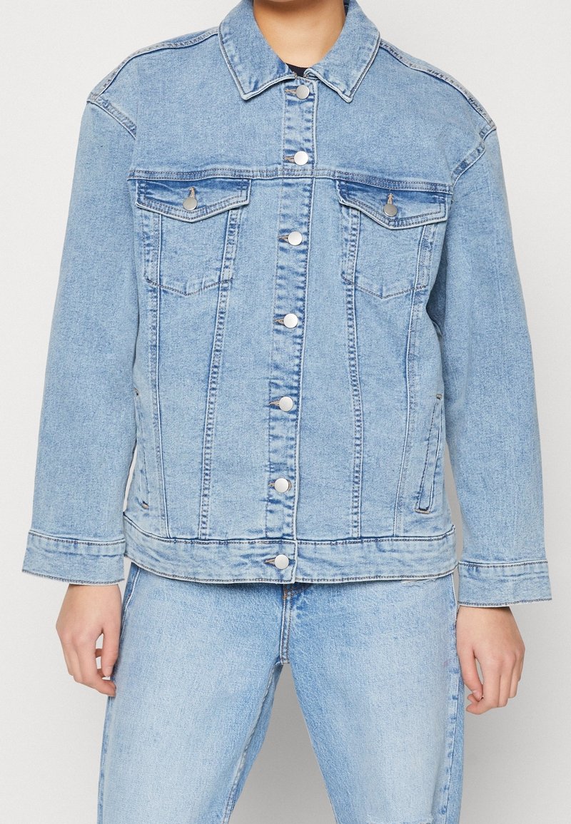 Person wearing light blue denim jacket with silver buttons and matching light blue denim jeans, standing against plain background.