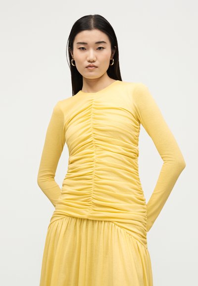 Jeune femme aux longs cheveux foncés, portant un haut jaune froncé à manches longues et une jupe assortie, debout devant un fond clair uni.