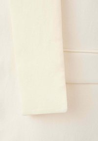 Manche d’un manteau en laine couleur crème avec un détail de couture près du poignet, présenté sur un fond en tissu crème assorti.