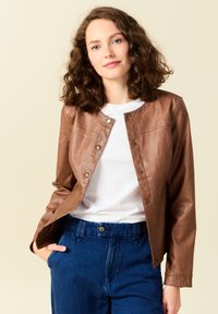 Giacca di pelle marrone con bottoni a pressione, colletto arrotondato e due tasche sul petto. Indossata sopra una camicia bianca, abbinata a pantaloni di jeans blu.