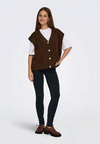 Νέα γυναίκα με λευκό κοντομάνικο πουκάμισο, καφέ γιλέκο με κουμπιά, σκούρα skinny τζιν και καφέ παπούτσια με κορδόνια, στέκεται με το χέρι στη γη.