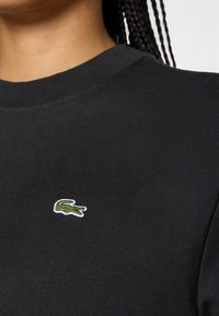 Gros plan d'une personne portant une chemise noire avec un petit logo crocodile vert brodé sur la poitrine.