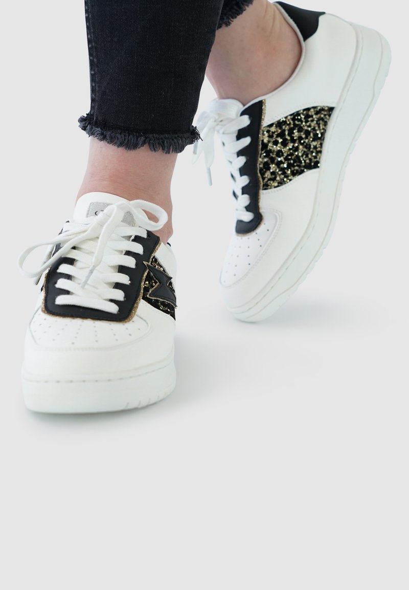 White and black sneakers featuring a glittery gold accent, smooth leather, and perforated detailing, paired with frayed black denim.