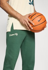 Krémový basketbalový dres s modrými číslami, doplnený zelenými teplákmi so zaobleným nápisom "Cream City" v bielej kurzíve. Hráč drží oranžový basketbal.