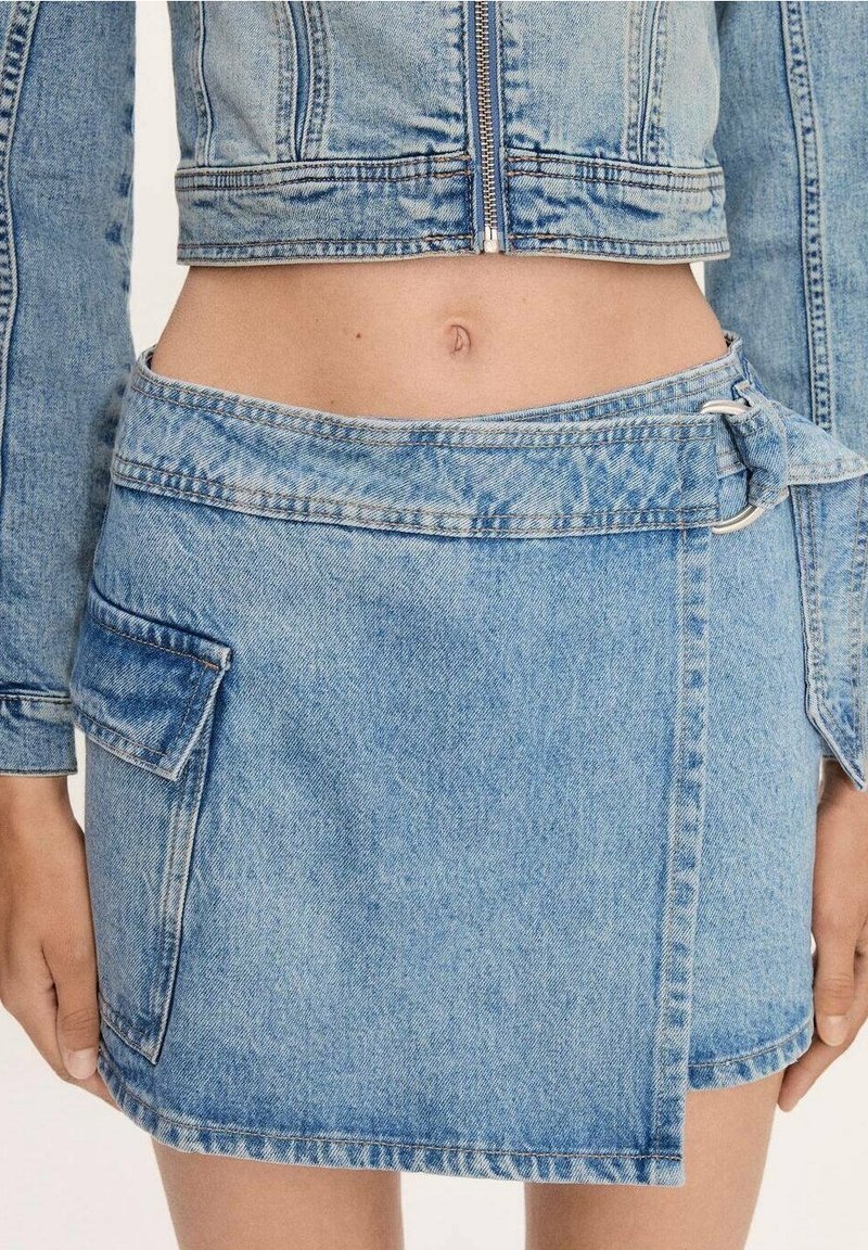 Vue rapprochée d'une personne portant une jupe en jean bleu clair avec une poche latérale et une veste en jean courte assortie avec une fermeture éclair.