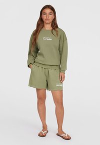 Completo felpa e pantaloni corti verde oliva, caratterizzato da una vestibilità rilassata e un logo in bianco. Il materiale sembra morbido con una texture liscia.