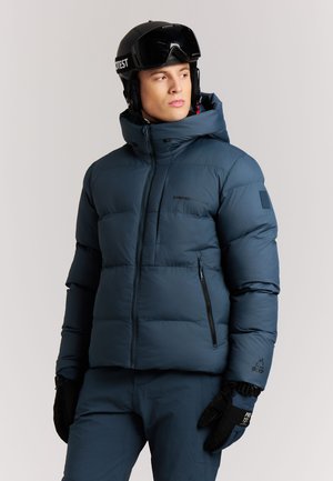 Man draagt een donkerblauwe gewatteerde winterjas, bijpassende broek, zwarte handschoenen en een zwarte skihelm met bril, kijkt naar opzij.