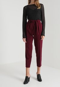 Maglietta nera a maniche lunghe con un motivo testurizzato, abbinata a pantaloni bordeaux a vita alta e mule neri, con un dettaglio della cintura annodata.