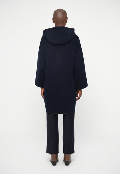 Cămașă cu glugă de culoare bleumarin, cu mâneci largi, dintr-un material texturat, tăiat drept, purtată peste pantaloni bleumarin asortați și pantofi cu toc maro.