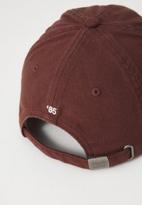 Căciulă bordeaux din material textil cu un finisaj texturat. Prezintă un mic broderie albă cu '85' și o curea metalică ajustabilă în spate.
