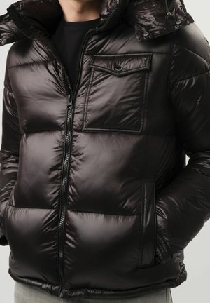 Mann trägt glänzende schwarze Steppjacke mit Reißverschluss vorne, Brustklappe, Seitentaschen und Kapuze über schwarzem Hemd.