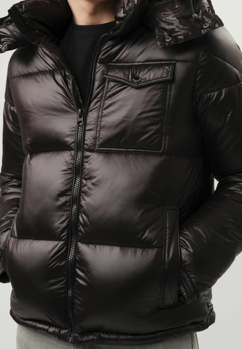 Homme portant une doudoune noire brillante avec fermeture à glissière à l'avant, poche poitrine à rabat, poches latérales et capuche, sur une chemise noire.