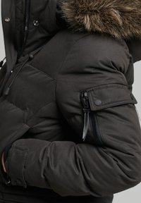 Czarna, ocieplana kurtka puchowa z kapturem wyłożonym sztucznym futrem. Do jej cech należy kieszeń zamykana na zamek na rękawie oraz ściągacze przy mankietach.