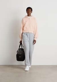 Felpa con zip rosa chiaro, pantaloni della tuta grigi, sneakers bianche e una borsa da palestra nera; tessuto morbido con vestibilità rilassata e polsini elastici.