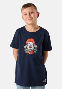 Navy puuvillatee, jossa on värikäs graafinen kuva koirasta, joka on pukeutunut punaiseen huppariin ja vihreisiin kuulokkeisiin. Rentoa tyyliä, lyhythihaisena.