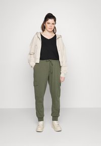 Cappotto corto beige, top nero a V e pantaloni cargo jogger verde oliva con tasche. Sneakers bianche completano l'outfit.