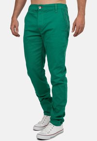 Chinos verdi in cotone a fit slim, con chiusura a bottone, dettagli in cucitura rossa e tasche laterali, abbinati a sneakers bianche.