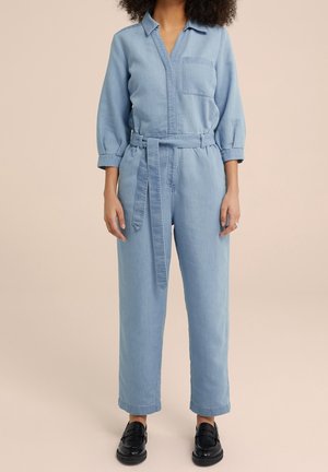 Kobieta ubrana w jasnoniebieski denimowy kombinezon z paskiem, przednią kieszenią na wysokości klatki piersiowej, rękawami do połowy łokcia oraz czarnymi butami wsuwanymi, stojąca na tle beżowego tła.