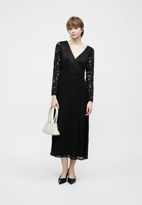 Kvinne iført en svart blonde langermet midi-kjole, svarte spisse leiligheter, holder en liten hvit håndveske, stående mot en hvit bakgrunn.