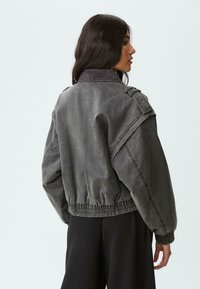 Grå denim bomberjakke med ribbet krave, elastisk kant og oversized skuldre. Har en let falmet tekstur og sidelommer.