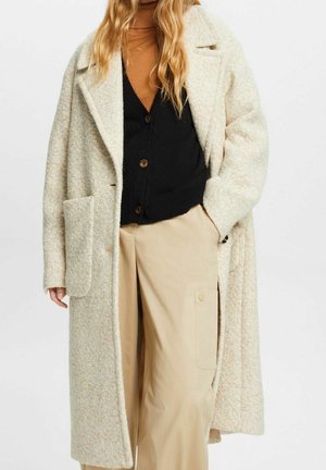 Femme portant un manteau en laine beige, un cardigan noir, un col roulé marron et un pantalon beige à jambes larges avec une poche latérale sur un fond uni.
