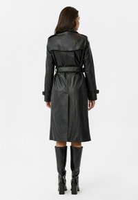 Zwarte leren trenchcoat met een ceintuur in de taille, lange mouwen en een lengte tot halverwege de kuit. Heeft een gladde textuur en een rugventilatie detail.