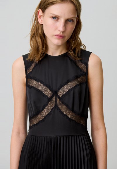 Vestido negro con falda plisada; el corpiño presenta detalles de encaje transparente en un patrón de cruzado; diseño sin mangas, textura de tela suave.