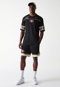 Black mesh sports jersey with gold and white stripes, featuring a logo on the chest. Matched with black shorts and white sneakers.