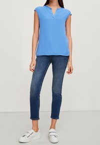 Hellblause Kurzarmbluse mit V-Ausschnitt, kombiniert mit dunkelblaue Skinny-Jeans und weißen Sneakers mit silbernen Hardware-Elementen.