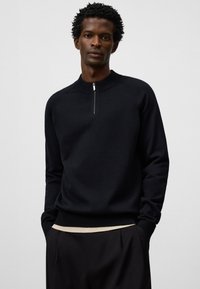 Svart stickad tröja med kvartslång dragkedja, ribbad krage och mudd, med en slät yta och ett kontrasterande beige band vid fållkanten.