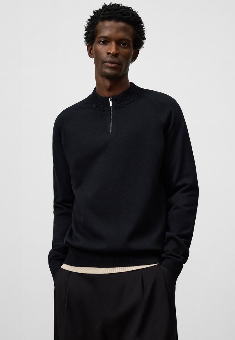 Svart stickad tröja med kvartslång dragkedja, ribbad krage och mudd, med en slät yta och ett kontrasterande beige band vid fållkanten.