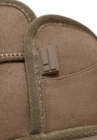 Parte superior de zapato de ante marrón con un acabado texturizado, que presenta una correa de nailon verde y un broche de plástico rectangular para el cierre.